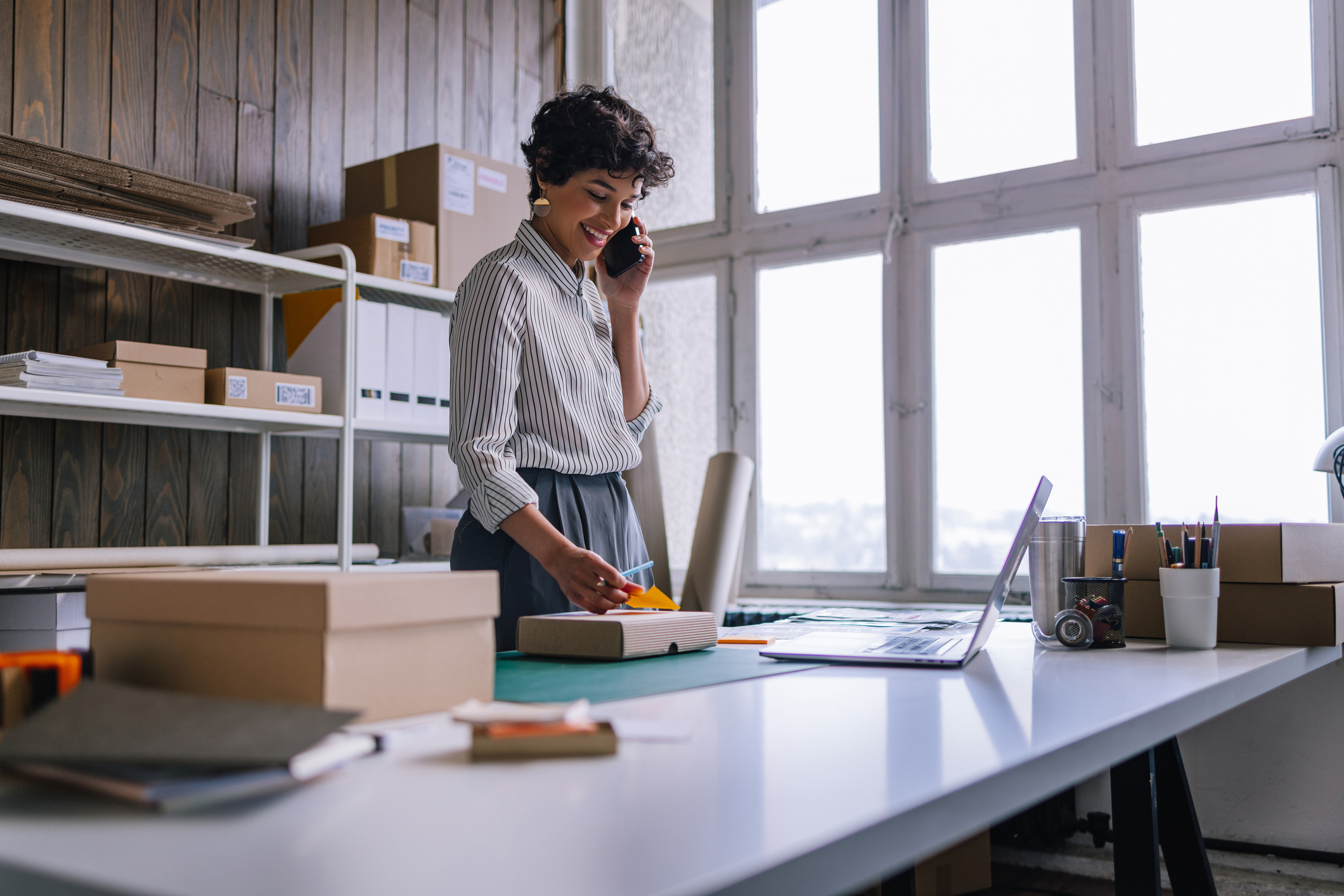 A Happy Businesswoman Talking On Her Smartphone While Preparing Packages For Shipping  In Her Store